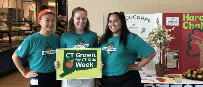 3 women holding a sign that says CT Grown for CT Kids week in front of a cafeteria's kitchen